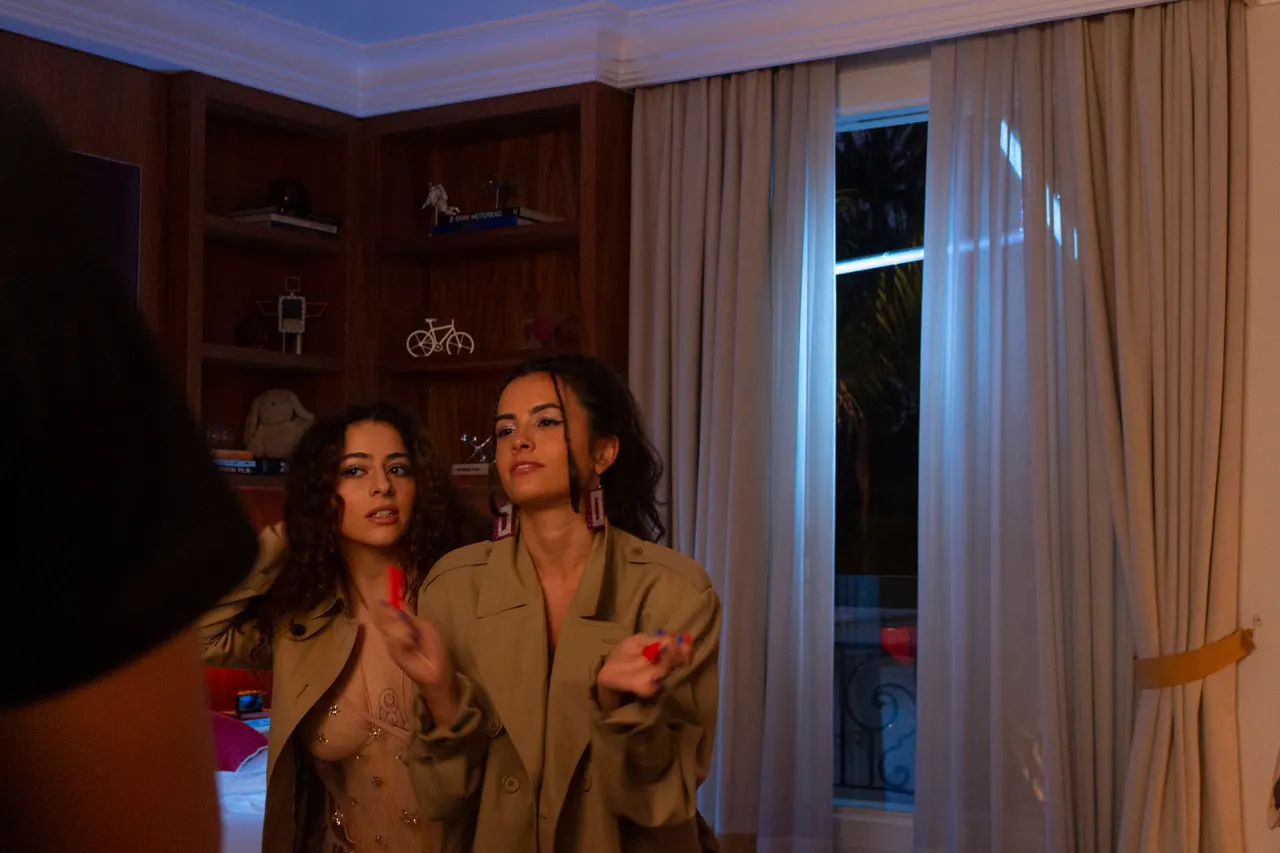Laura e a amiga de trench coat por cima de roupas brilhantes, junto à janela do quarto.