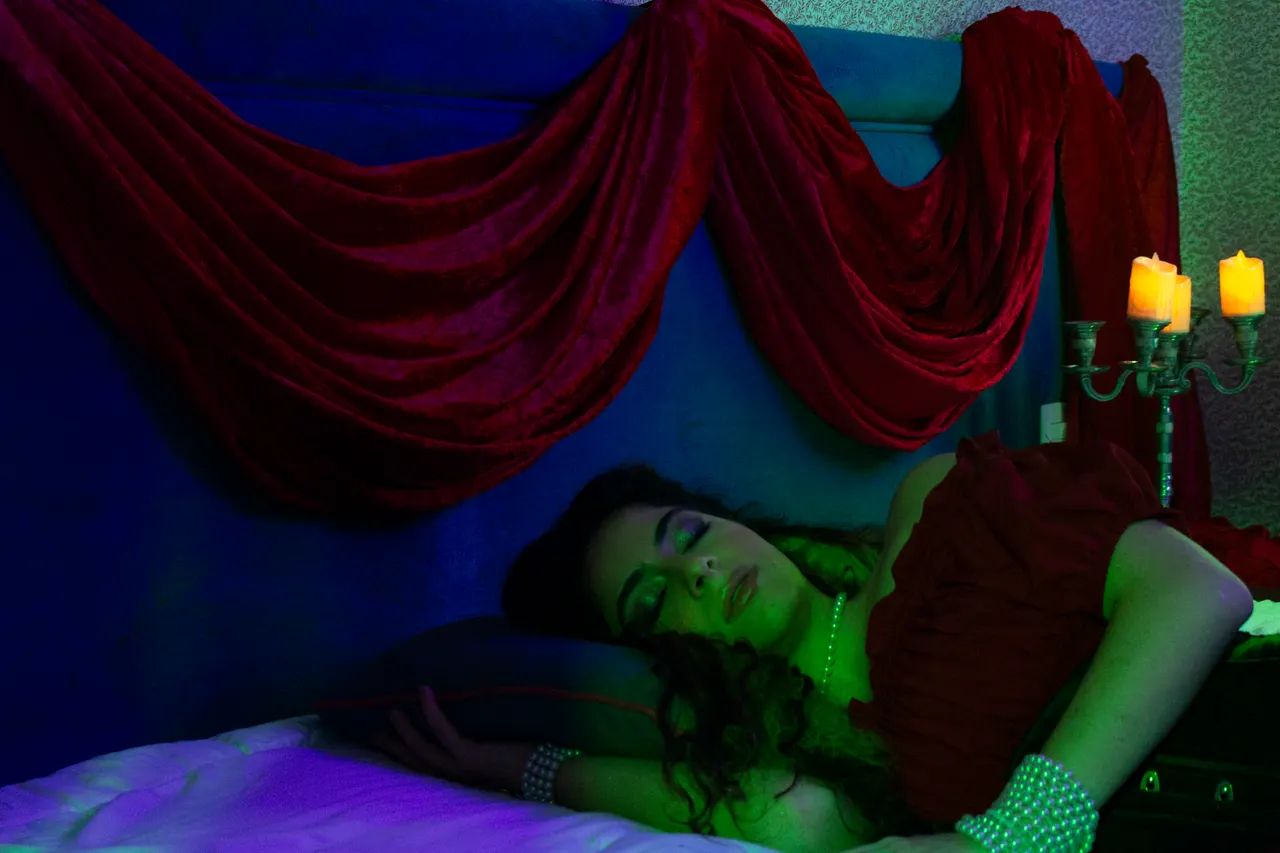 Laura, de vestido vermelho, deitada sob cortinas de veludo vermelho, com velas ao lado.