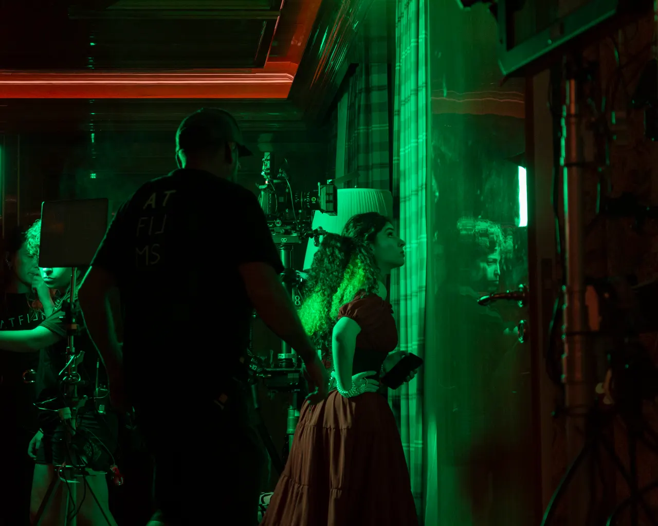 A equipe filma Laura de vestido vermelho junto a uma janela, com a sala em verde.