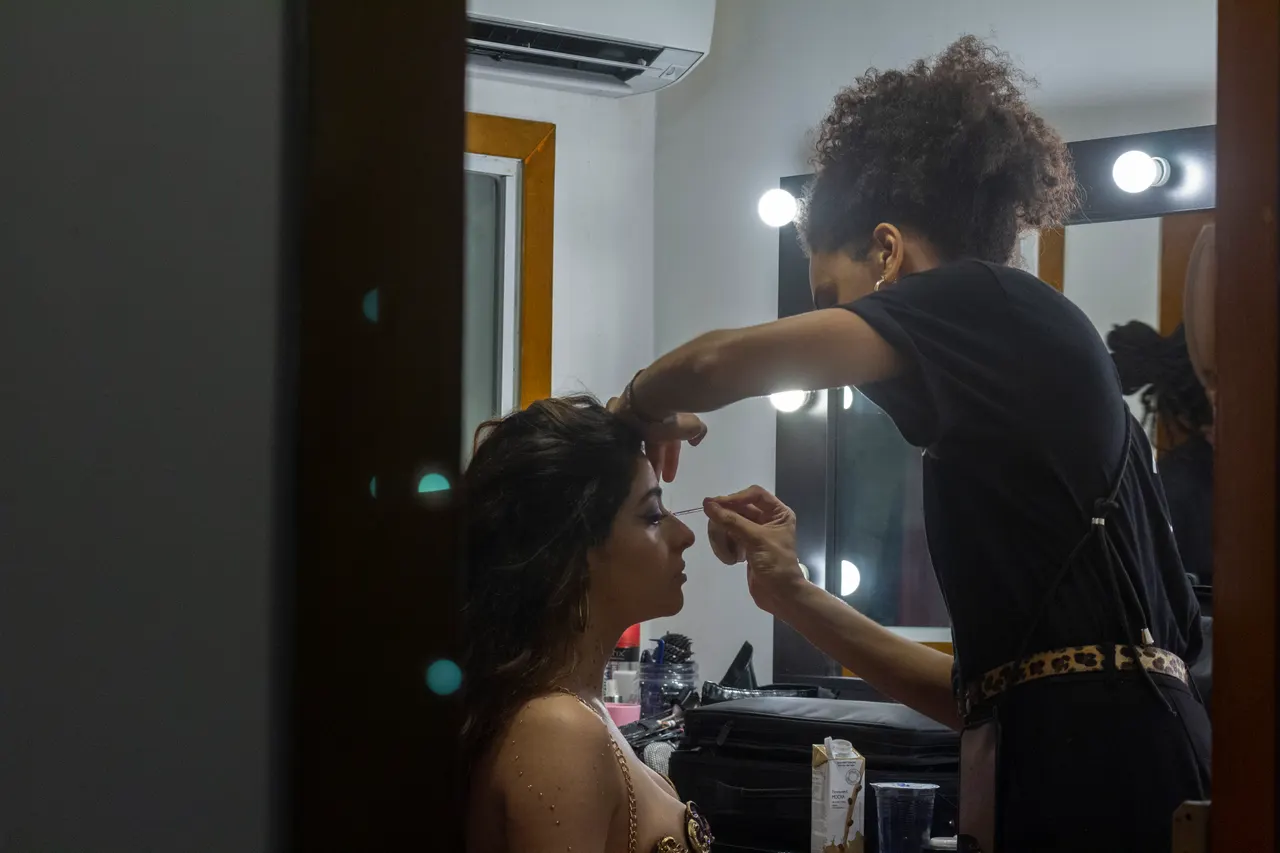 Uma maquiadora trabalha nos cílios de Laura diante do espelho de camarim.