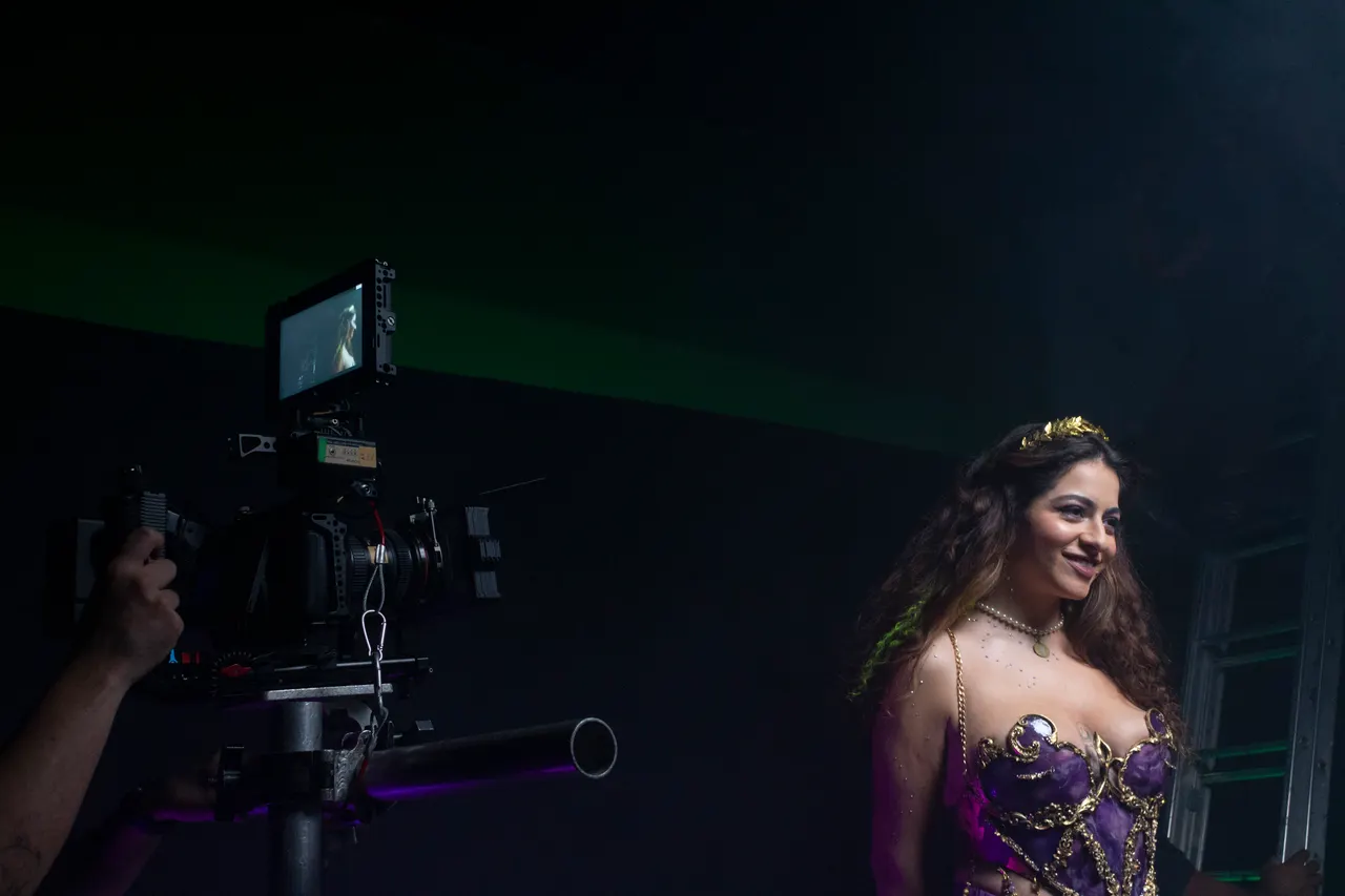 Laura, de corpete roxo e dourado e coroa, sorri para a câmera estabilizada na fumaça.