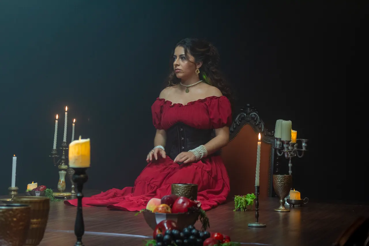 Laura, de vestido vermelho e espartilho preto, ajoelhada na mesa, com pérolas nos pulsos.
