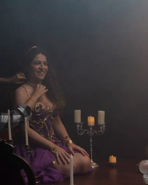 Mãos ajustam o colar de Laura, ajoelhada no figurino roxo.